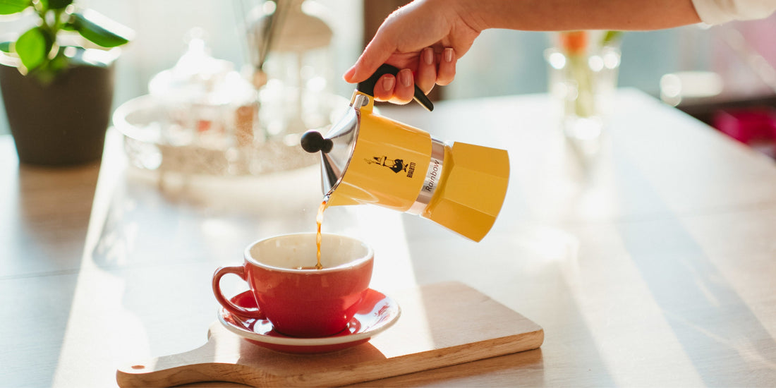 Hand pouring fresh coffee from yellow moka pot into red cup on sunny kitchen table