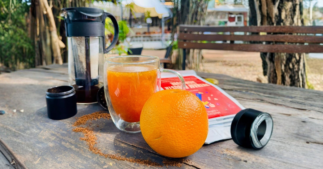Glass of orange coffee, fresh orange, coffee maker, and coffee grounds on wooden table outdoors.