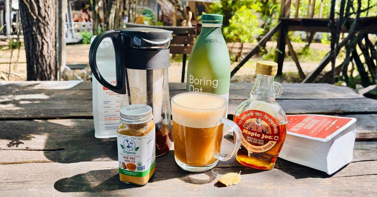 Outdoor table with oat milk, French press coffee, cinnamon, maple syrup, and a glass of frothy coffee.