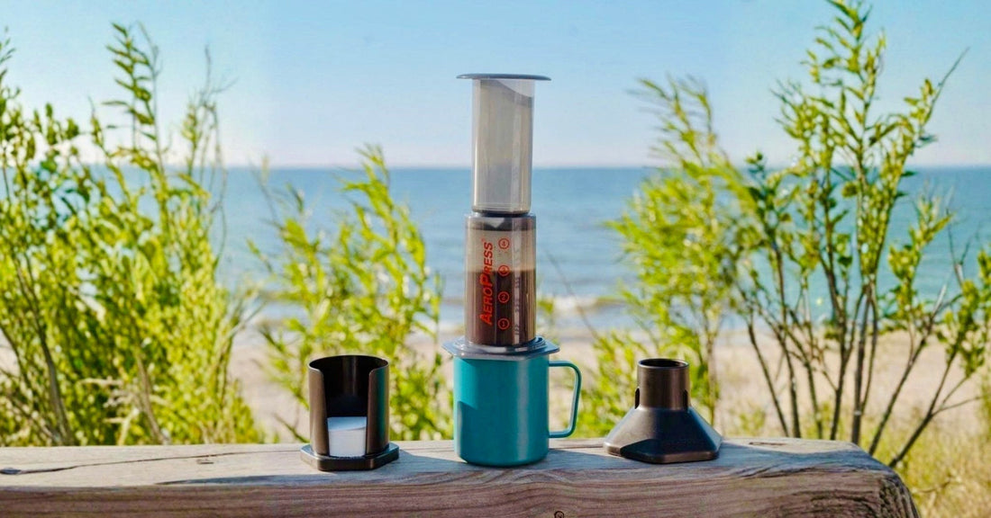 Aeropress coffee maker with blue mug on wooden ledge outdoors by the beach, summer morning scene
