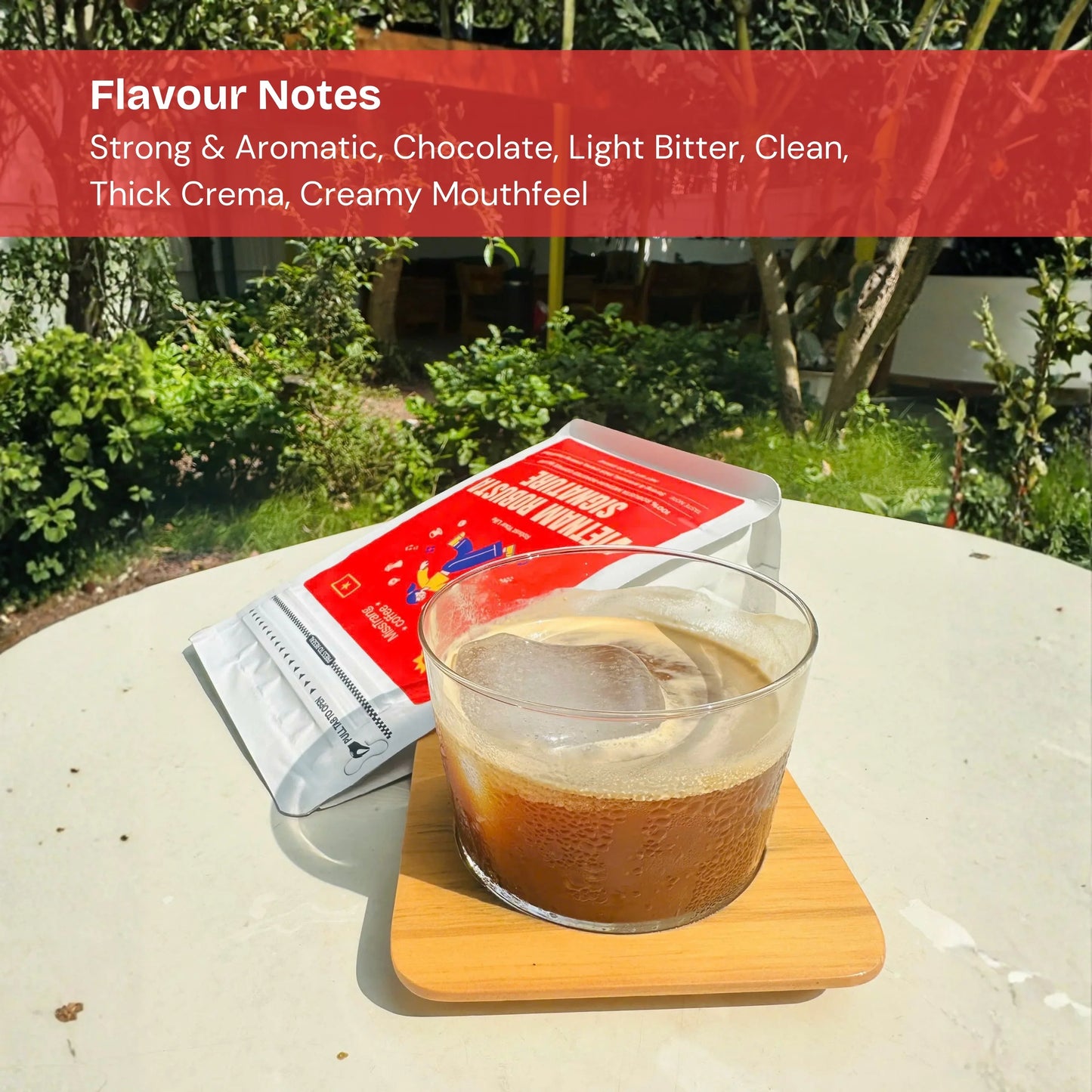Iced coffee with thick crema in a clear glass on a wooden coaster, coffee bag in background, outdoor garden setting