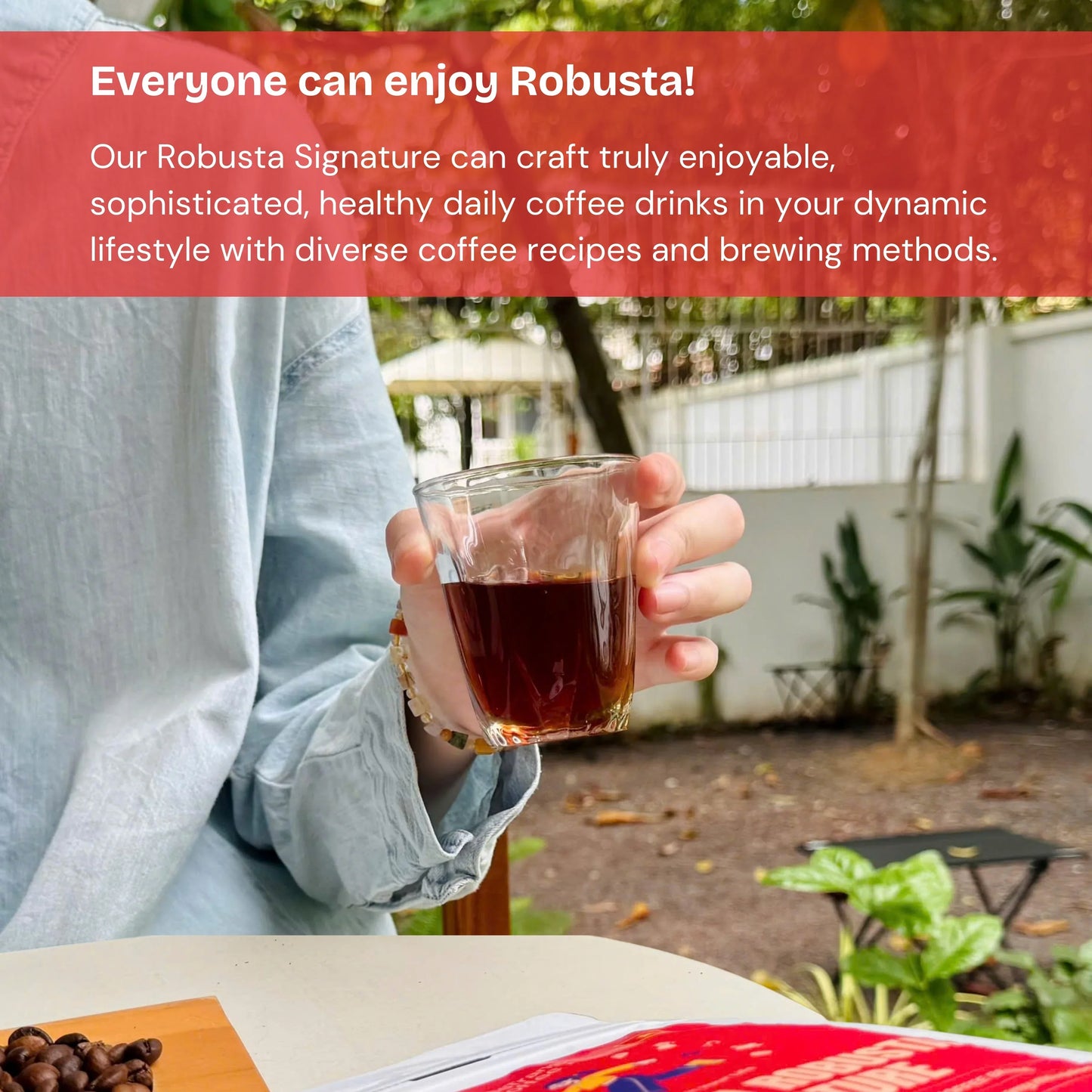 Hand holding a glass of black Robusta coffee outdoors, with coffee beans and greenery nearby.