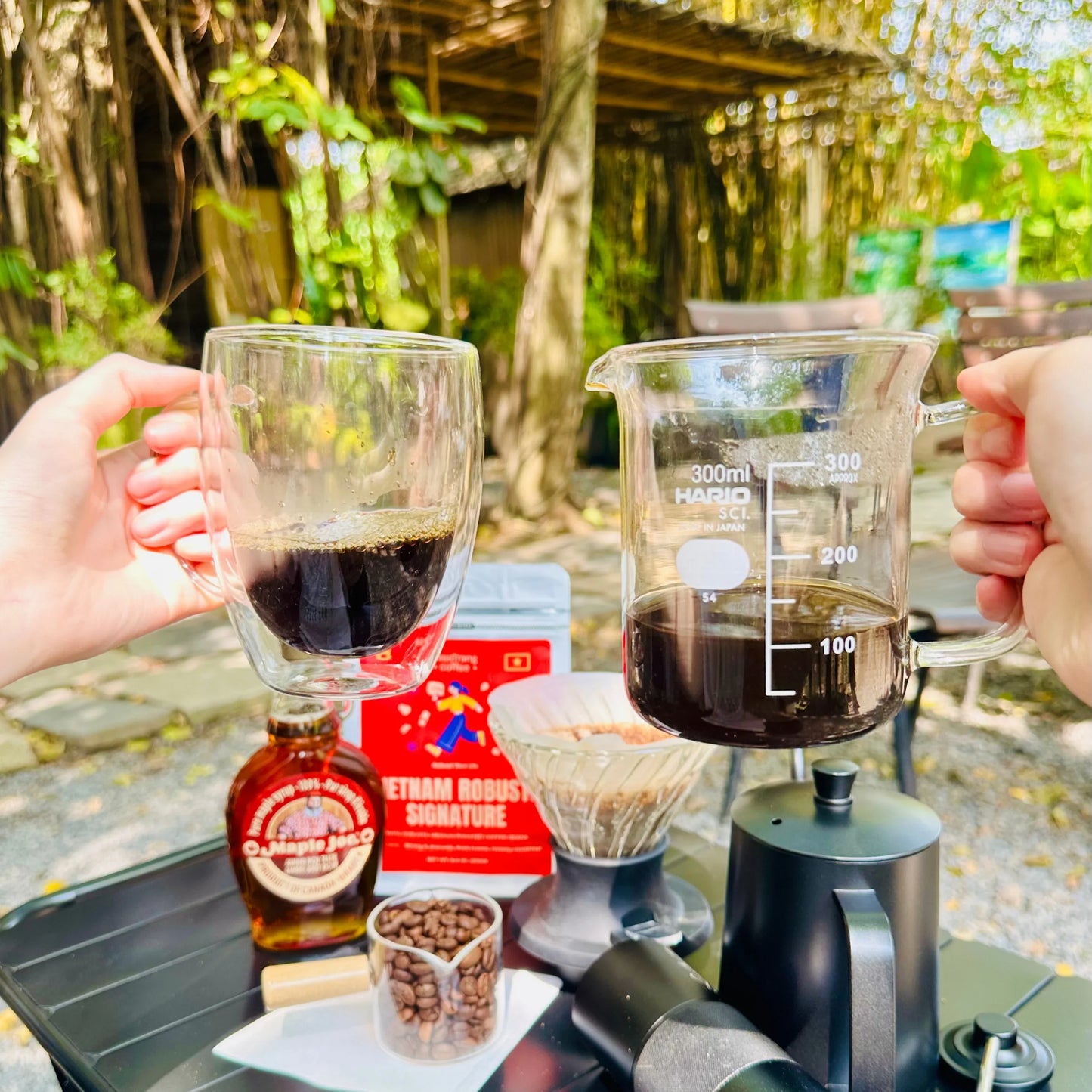Outdoor coffee setup with glass mug, beaker of Vietnamese coffee, coffee beans, and pour-over kit